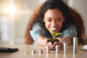 Young happy hispanic woman holding a plant growing out of soil in her hands with money coins on different levels on a table. Symbolizes money growing, investment and banking